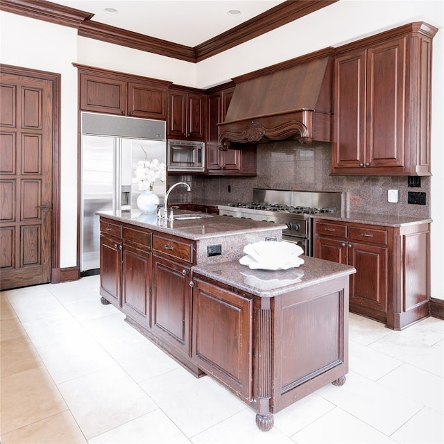 kitchen featuring stone counters, premium range hood, sink, built in appliances, and a center island with sink