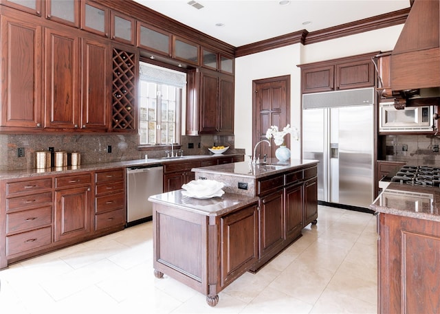 kitchen featuring decorative backsplash, built in appliances, crown molding, custom range hood, and a center island with sink