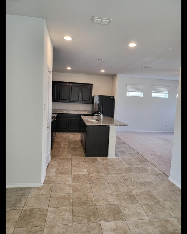 kitchen featuring visible vents, an island with sink, light stone counters, freestanding refrigerator, and a sink