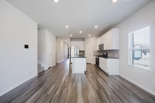 kitchen featuring dark wood-style floors, an island with sink, stainless steel appliances, dark countertops, and tasteful backsplash