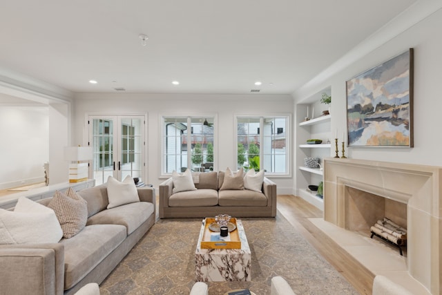 living room with a fireplace, crown molding, built in shelves, light wood-type flooring, and french doors