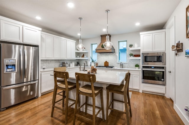 kitchen featuring decorative light fixtures, white cabinetry, an island with sink, stainless steel appliances, and custom range hood