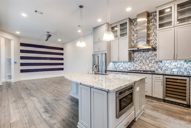 kitchen featuring pendant lighting, wall chimney range hood, appliances with stainless steel finishes, a kitchen island with sink, and beverage cooler