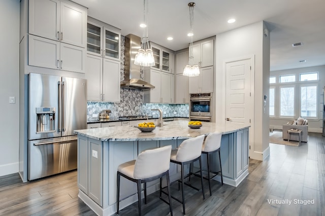 kitchen featuring wall chimney range hood, a center island with sink, light stone countertops, and appliances with stainless steel finishes