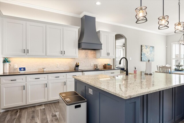 kitchen featuring dark stone counters, custom range hood, decorative light fixtures, white cabinetry, and a sink