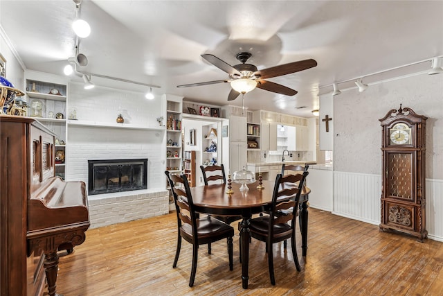 dining area with a wainscoted wall, ornamental molding, a ceiling fan, a brick fireplace, and wood finished floors