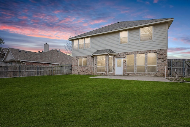 back of property at dusk featuring a lawn, a fenced backyard, a shingled roof, brick siding, and a patio area