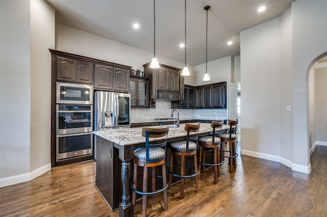 kitchen with appliances with stainless steel finishes, hanging light fixtures, a kitchen island with sink, light stone countertops, and dark brown cabinets