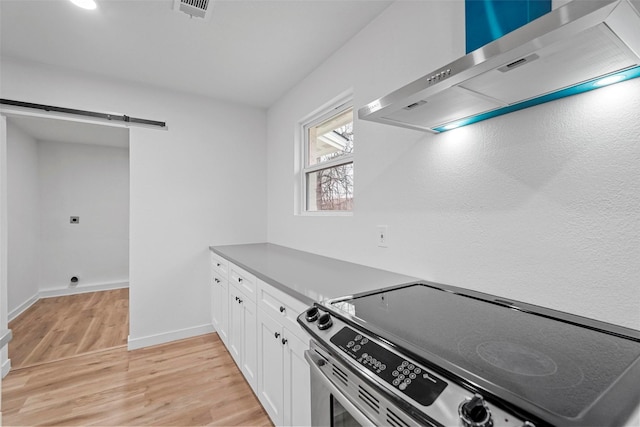 kitchen with wall chimney range hood, stainless steel electric range, white cabinets, a barn door, and light wood-type flooring