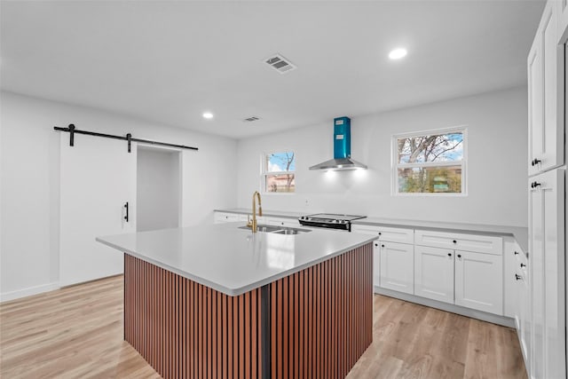 kitchen featuring wall chimney exhaust hood, sink, white cabinetry, an island with sink, and a barn door