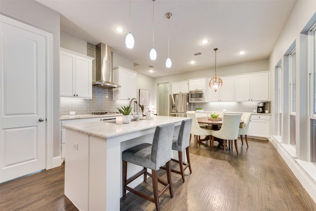 kitchen with wall chimney range hood, white cabinetry, stainless steel appliances, an island with sink, and decorative light fixtures