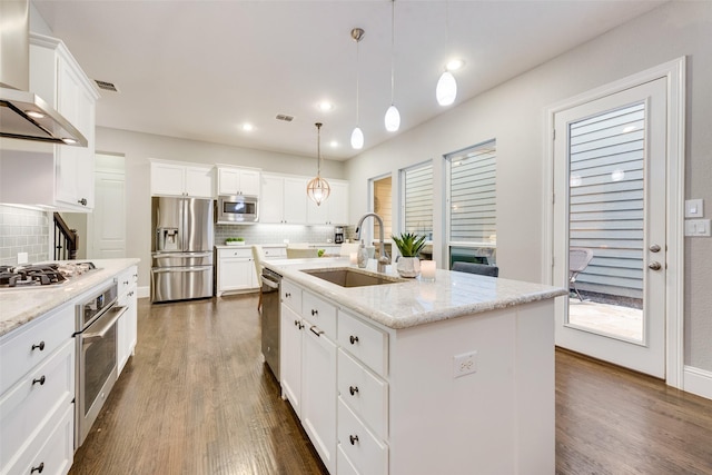 kitchen with wall chimney range hood, appliances with stainless steel finishes, a kitchen island with sink, white cabinetry, and hanging light fixtures