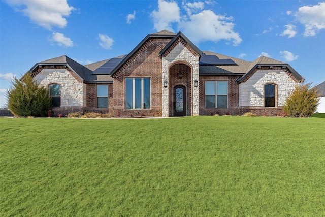 french country home with solar panels, brick siding, a shingled roof, stone siding, and a front lawn