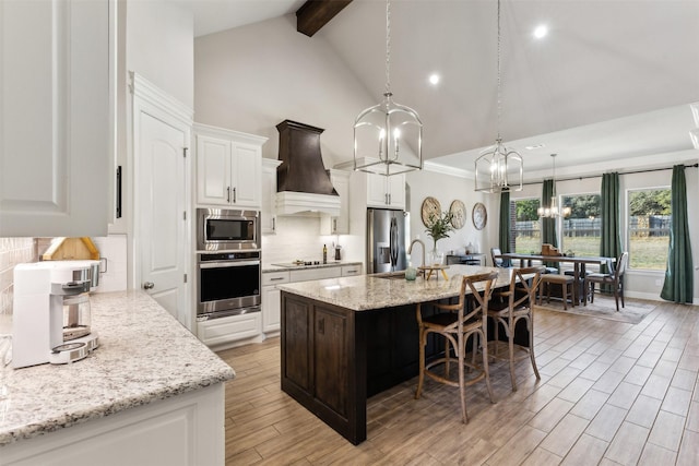 kitchen featuring a center island with sink, custom range hood, appliances with stainless steel finishes, light stone counters, and white cabinetry