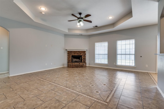 unfurnished living room with a tray ceiling, ceiling fan, light tile patterned floors, and a brick fireplace