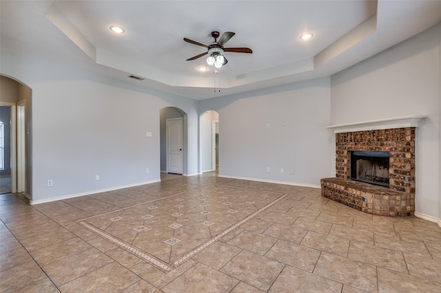 unfurnished living room featuring a tray ceiling, ceiling fan, light tile patterned floors, and a brick fireplace