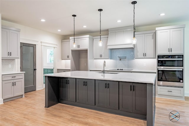 kitchen with sink, light wood-type flooring, custom range hood, black electric stovetop, and a kitchen island with sink