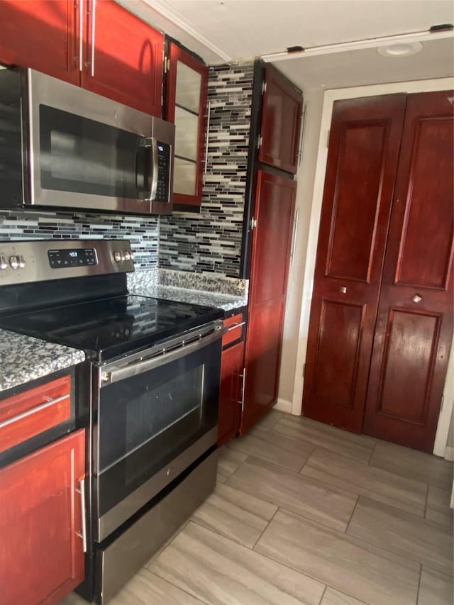 kitchen featuring reddish brown cabinets, backsplash, appliances with stainless steel finishes, stone countertops, and glass insert cabinets