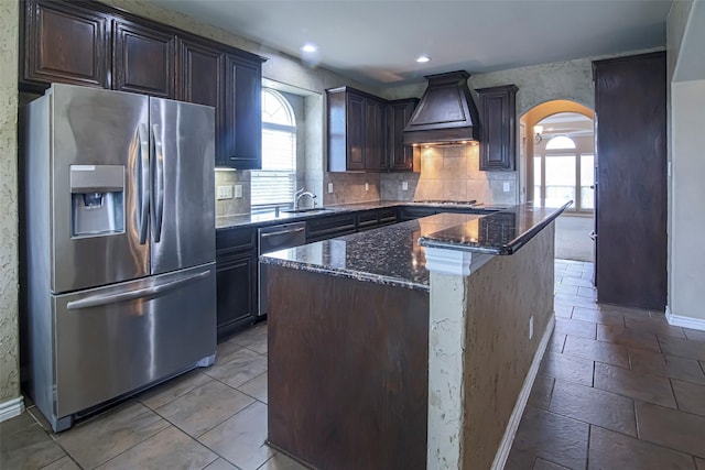 kitchen with dark brown cabinets, stainless steel appliances, a center island, custom exhaust hood, and dark stone counters