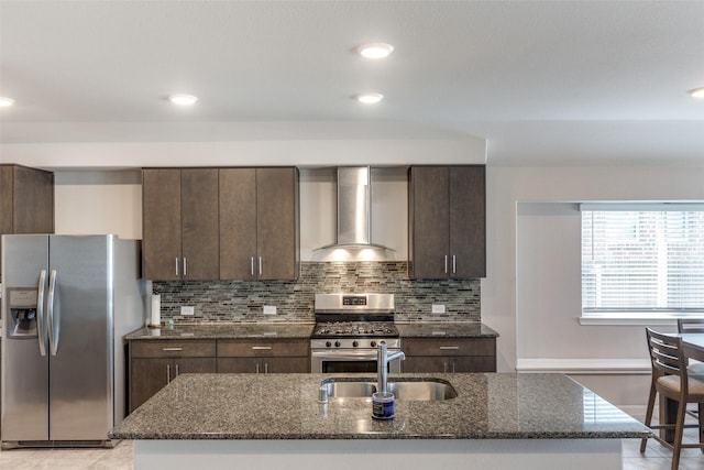 kitchen featuring appliances with stainless steel finishes, sink, dark stone counters, a kitchen island with sink, and wall chimney exhaust hood