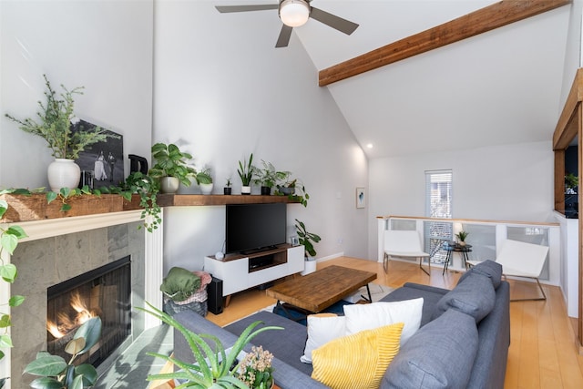 living room featuring high vaulted ceiling, beamed ceiling, a tiled fireplace, ceiling fan, and light hardwood / wood-style floors