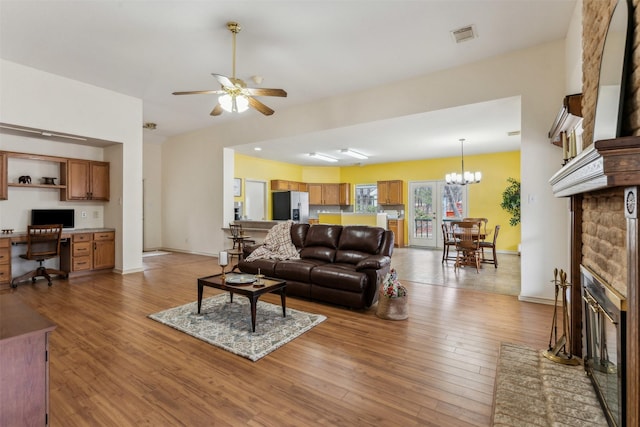 living area with built in study area, ceiling fan with notable chandelier, wood finished floors, and a stone fireplace