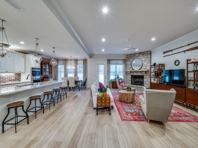 living room with a stone fireplace, ceiling fan with notable chandelier, sink, ornamental molding, and light hardwood / wood-style floors