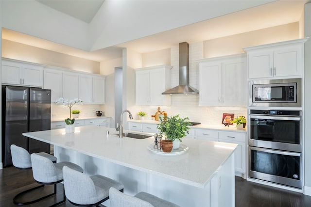 kitchen with appliances with stainless steel finishes, an island with sink, sink, white cabinets, and wall chimney range hood