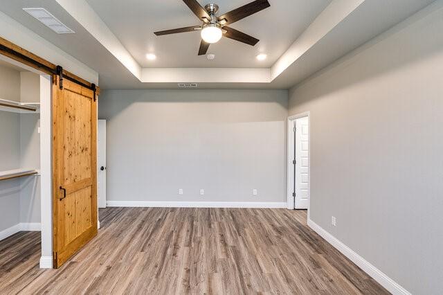 unfurnished bedroom with a tray ceiling, wood-type flooring, a barn door, and ceiling fan