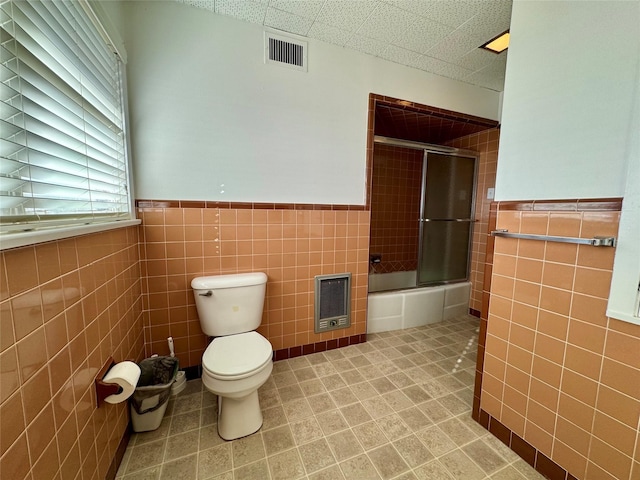 bathroom featuring combined bath / shower with glass door, heating unit, tile walls, and toilet