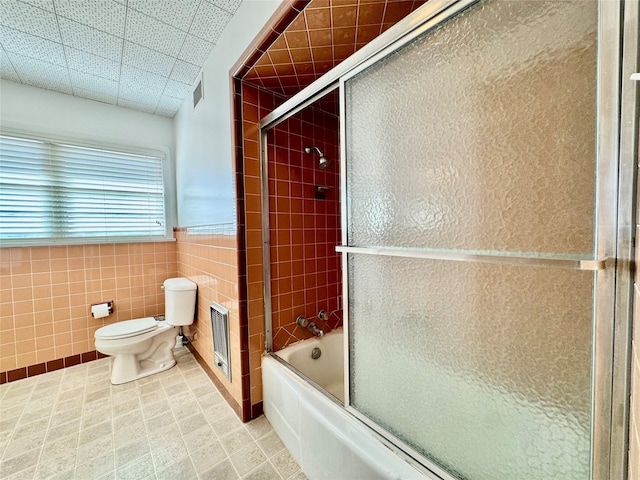 bathroom with heating unit, tile walls, shower / bath combination with glass door, and toilet