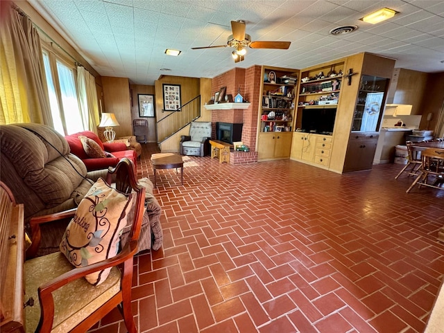 living room featuring ceiling fan, a brick fireplace, and wooden walls