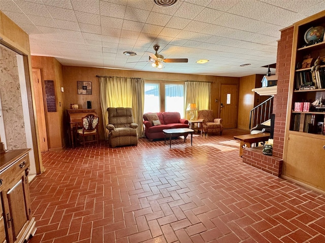 living area featuring ceiling fan, a brick fireplace, and wood walls