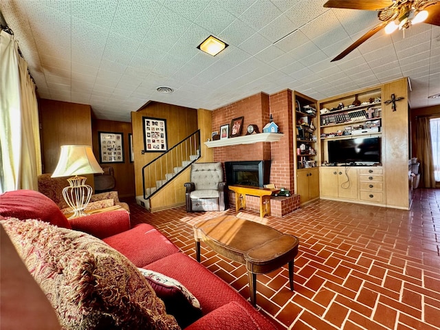 living room featuring built in shelves, a brick fireplace, ceiling fan, and wood walls