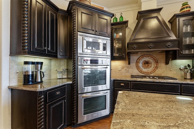 kitchen with premium range hood, backsplash, stainless steel appliances, light stone countertops, and ornamental molding