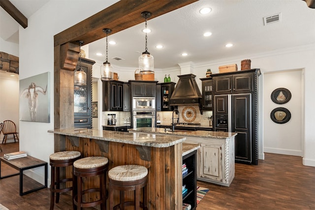 kitchen with built in appliances, custom range hood, pendant lighting, light stone countertops, and a kitchen island with sink