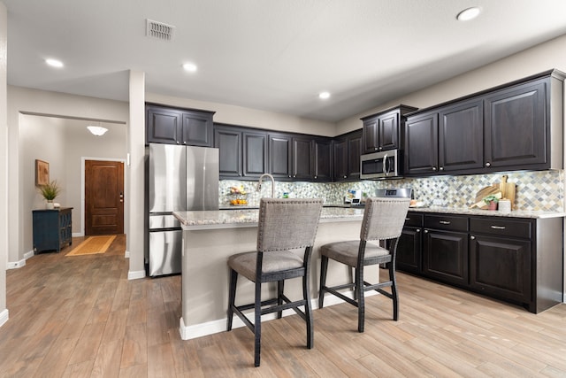 kitchen featuring a breakfast bar, light wood-type flooring, an island with sink, stainless steel appliances, and light stone countertops