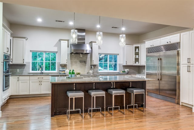 kitchen featuring wall chimney exhaust hood, white cabinetry, hanging light fixtures, appliances with stainless steel finishes, and a kitchen island with sink