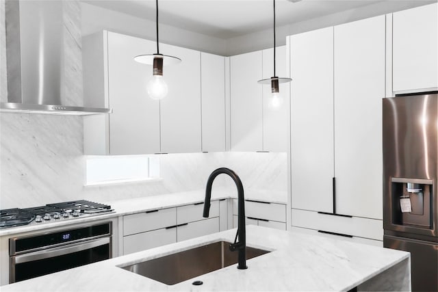 kitchen with pendant lighting, wall chimney range hood, sink, white cabinetry, and stainless steel appliances