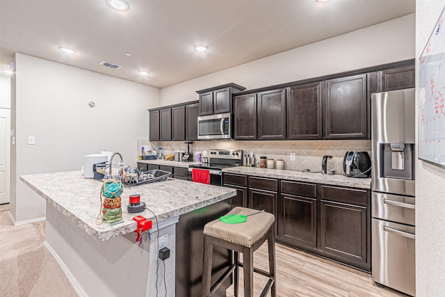 kitchen with appliances with stainless steel finishes, an island with sink, a breakfast bar area, backsplash, and dark brown cabinetry