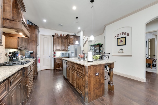 kitchen with pendant lighting, custom exhaust hood, light stone counters, stainless steel appliances, and a center island with sink