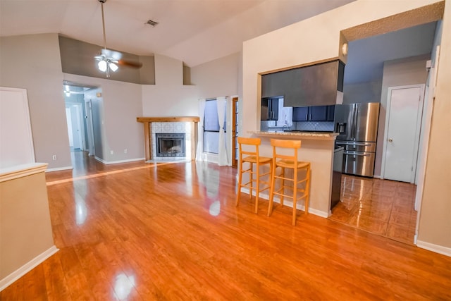 kitchen featuring a breakfast bar, stainless steel fridge, ceiling fan, a tiled fireplace, and light hardwood / wood-style floors