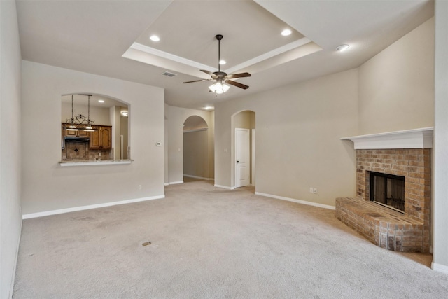 unfurnished living room featuring ceiling fan, a tray ceiling, light colored carpet, and a brick fireplace