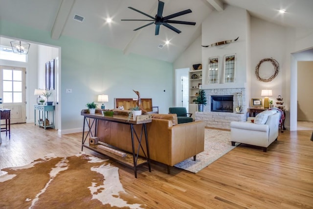 living room with a stone fireplace, ceiling fan with notable chandelier, high vaulted ceiling, light hardwood / wood-style floors, and beam ceiling