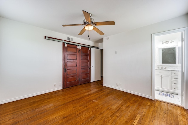 unfurnished bedroom featuring connected bathroom, sink, wood-type flooring, ceiling fan, and a barn door