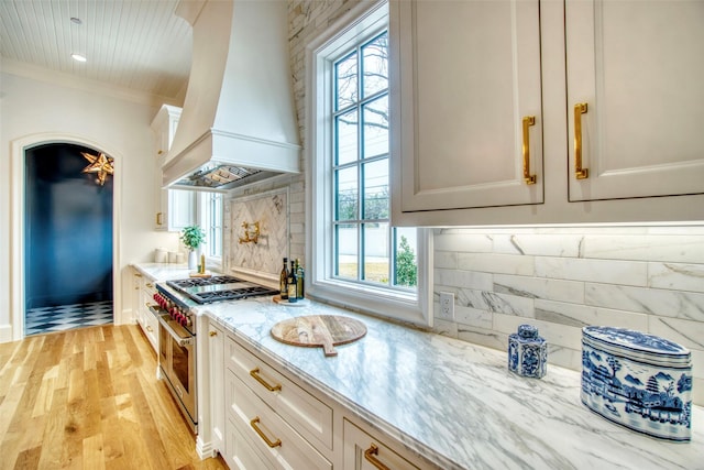 kitchen featuring light wood finished floors, range with two ovens, custom range hood, light stone countertops, and backsplash