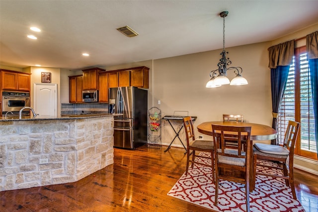 kitchen with dark wood-type flooring, backsplash, hanging light fixtures, stainless steel appliances, and dark stone counters