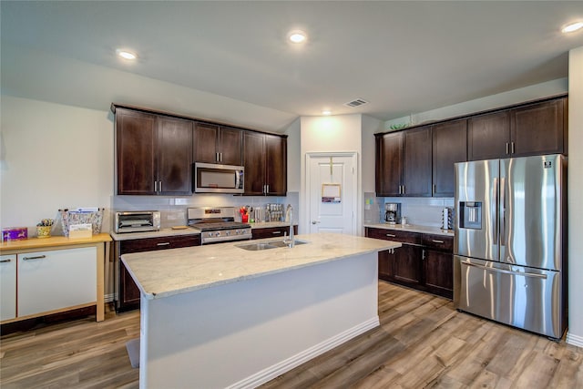 kitchen with sink, wood-type flooring, dark brown cabinets, an island with sink, and stainless steel appliances