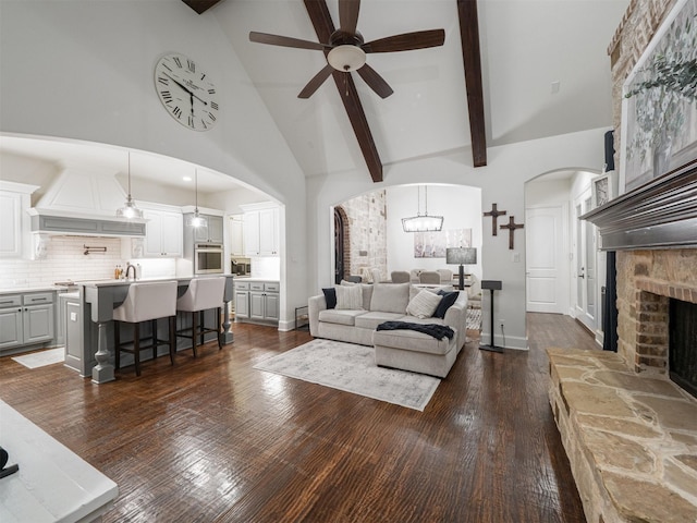 living room with ceiling fan, high vaulted ceiling, dark hardwood / wood-style floors, a fireplace, and beamed ceiling