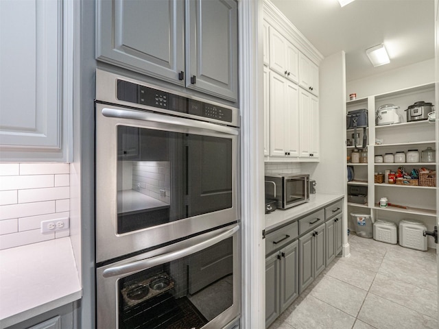 kitchen with gray cabinetry, light tile patterned floors, white cabinets, stainless steel double oven, and backsplash